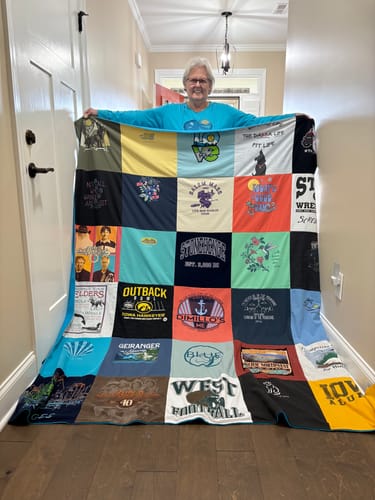 Customer holding up a large, colorful Premium T Shirt Quilt made from various t-shirts in a hallway.