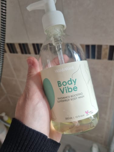 Reviewer holding the clear pump bottle of Body Vibe body wash in a shower.
