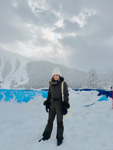 Reviewer in a dark brown one-piece ski suit with faux fur cuffs, standing on a snowy mountain.