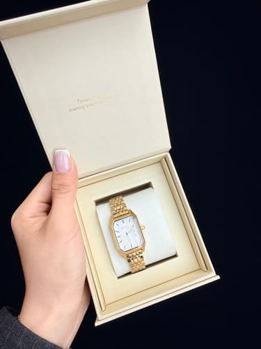 Customer's hand holding open a cream-colored box to show the Isabell Gold watch with its white, rectangular face.
