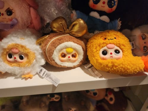 Reviewer displays three adorable Baby Three Big Bite plush toys, including the secret character with a gold bow, on a shelf.