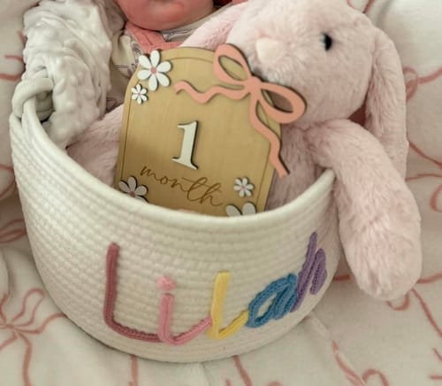Customer's white knitted basket with a colorful personalized name, holding a baby, a stuffed bunny, and a 1-month sign.