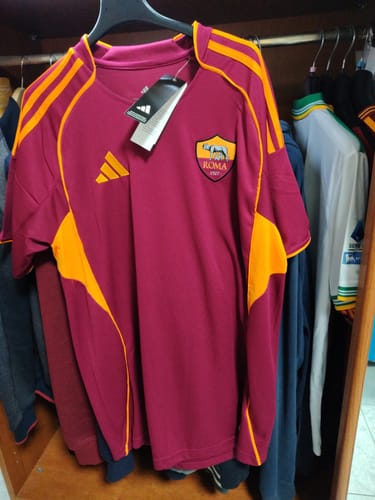 Un recensore mostra la Maglia Roma Home amaranto con dettagli arancioni, appesa su una gruccia.