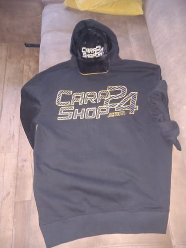 Beoordelaar toont een zwarte, fluwelen CARPSHOP24-pet boven op een hoodie.