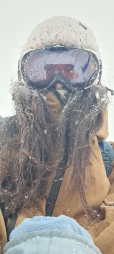 Close-up of a reviewer wearing the caramel snow jacket in heavy snowfall, with snow covering their hair and goggles.