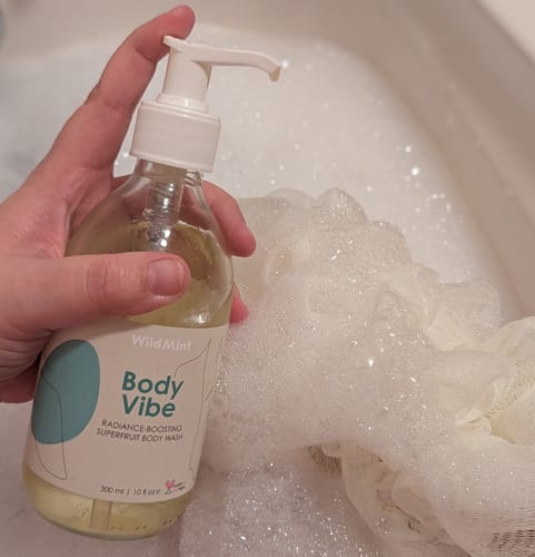 Customer holding a bottle of Body Vibe body wash next to a sudsy sponge in a bath.