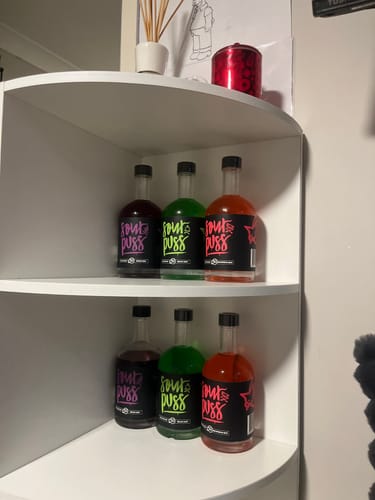 Customer's Sour Puss bottles in grape, apple, and watermelon flavors organized on two white shelves.