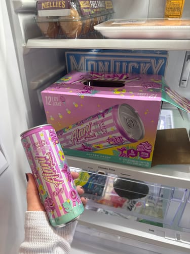 Customer holds a colorful Alani Nu Energy Drink can in front of an open fridge with a 12-pack case inside.