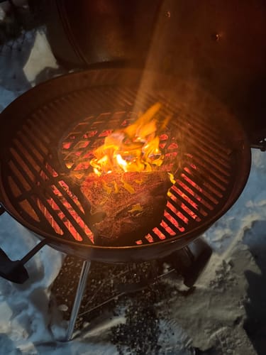Customer grilling a thick steak over hot coals on a kettle grill set in the snow.