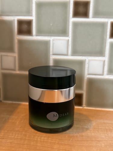 レビューアーのPG クリーミィリッチセラム。タイル壁の前の木製カウンターに置かれた深緑の容器。