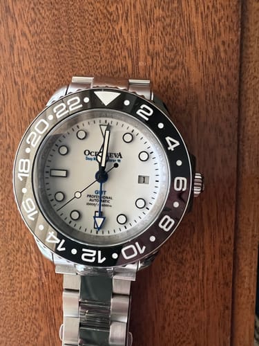 Customer's Oceaneva GMT watch with a white dial, black bezel, and a steel bracelet on a wood surface.