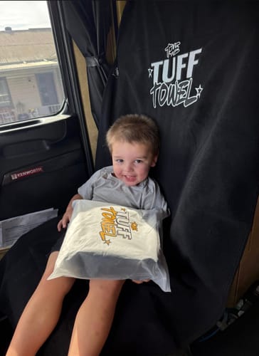 Reviewer's child sits on the black Waterproof Car Towel Seat Cover in a truck, holding the product's packaging.
