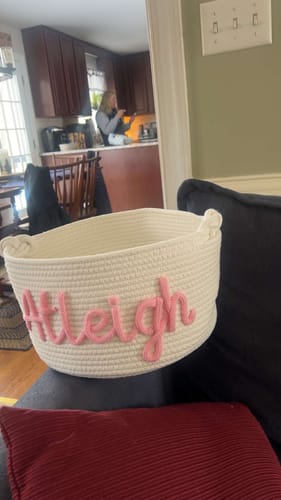 Customer's personalized white woven basket with the name 'Atleigh' in pink, resting on a dark piece of furniture.