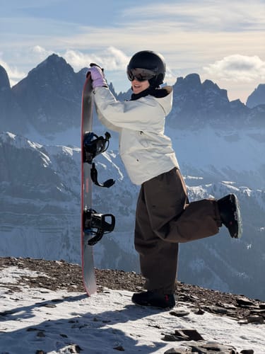 Reviewer on a snowy mountain wearing the baggy coffee snow pants while holding a snowboard.