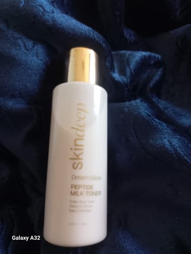 Customer's white bottle of DREAM GLAZE Peptide Milk Toner with a gold cap, set against a dark blue textured fabric.