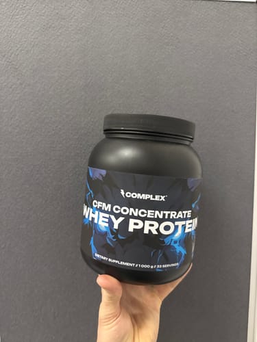 A reviewer holds a large black tub of Complex CFM Concentrate Whey Protein against a gray background.