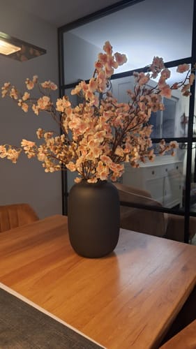 Klant toont de oudroze Zijden Bloesem in een donkere vaas, mooi geplaatst op een houten eettafel.