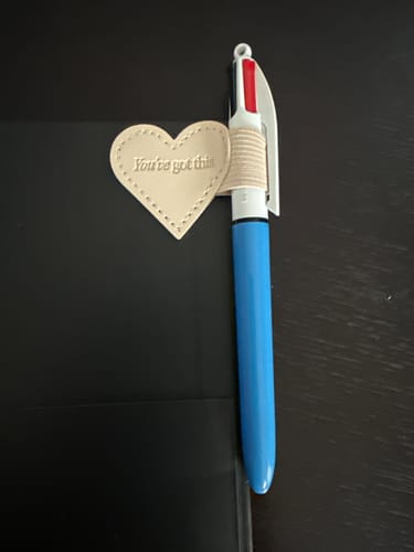 Customer's beige heart-shaped Stick-on Pen Loop with the quote 'You've got this' attached to a black planner, holding a pen.