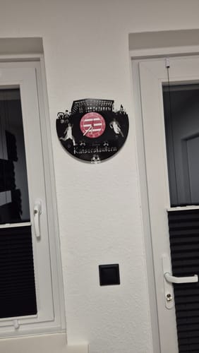 Kunde zeigt die Fussball-Wanduhr 'Kaiserslautern' aus Vinyl, die an einer weißen Wand hängt.