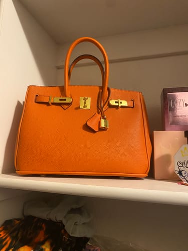 Customer's bright orange LALUVA Warkin Bag with gold hardware, sitting on a white closet shelf.