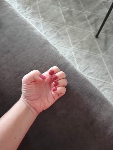 Hand einer Rezensentin zur Faust geballt, die die 'Grapevine Twist' UV-Gel-Nagelfolien in einem schönen Pflaumenrot auf kurzen Nägeln zeigt.