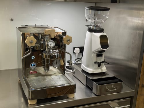 Reviewer's white Fiorenzato Allground Coffee Grinder on a stainless steel counter next to a chrome espresso machine.