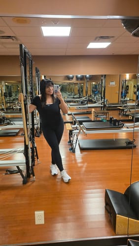 Reviewer standing in a pilates studio wearing the full-length black The Onesie with cream straps.