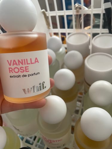 Reviewer holding a frosted glass bottle of Vanilla Rose perfume with other fragrances in a basket behind it.