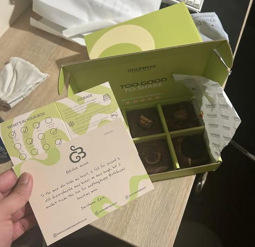 Reviewer holding a gift card next to an open Happy Birthday Mixed Box of Postal Brownies with four assorted treats inside.