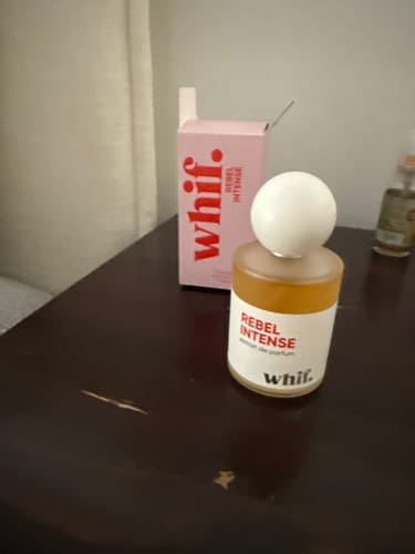 Customer's bottle of Rebel Intense perfume next to its pink box on a dark wooden table.