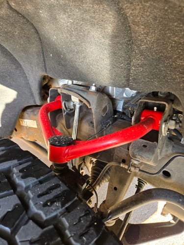 Reviewer's installed bright red upper control arm viewed from above, next to a large tire.