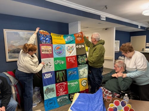 Reviewer's family presenting a large, colorful Premium T Shirt Quilt to a seated man who is visibly moved.