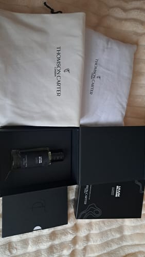 Customer's unboxed Fragrance & Body Wash Bundle displaying the bottle in a black box alongside two branded white cloth pouches.