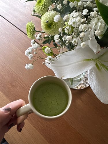 Client tenant une tasse de Thé Matcha Premium Bio vert et mousseux sur une table en bois.