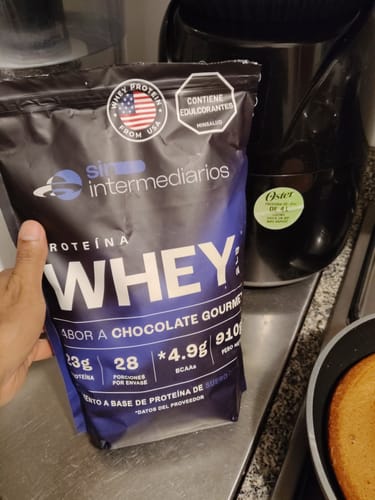Un cliente sostiene la bolsa de Proteína Whey - Chocolate en una cocina, junto a un sartén con una torta hecha con el producto.