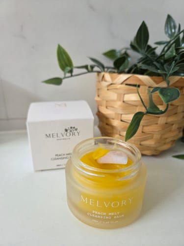 Customer's open jar of golden Peach Melt Cleansing Balm with a rose quartz spatula resting inside.