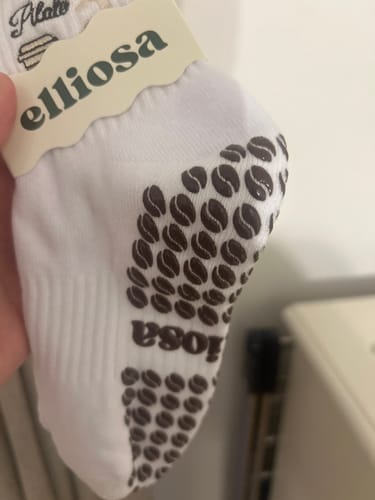 Customer showing a close-up of a white Pilates & Lattes sock, highlighting the brown, coffee bean-shaped grips on the sole.