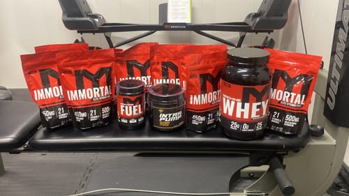 Reviewer's red Immortal Elite Multi-Vitamin Packs displayed with other supplements on a workout bench.