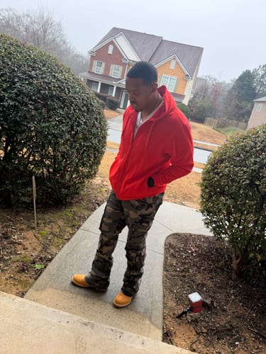 Customer standing on a doorstep wearing a red hoodie and camouflage pants, looking down.
