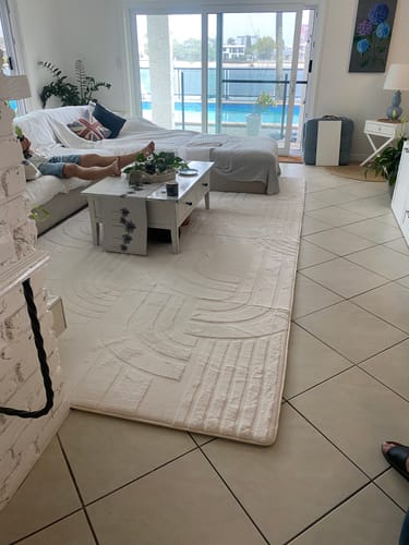 Customer's large, cream Mellow Mat laid out in a bright living room, with a person relaxing on the sofa in the background.