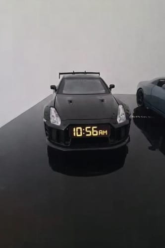 Reviewer's black Hypercar Alarm Clock on a desk, displaying "10:56 AM" on its front LED screen.