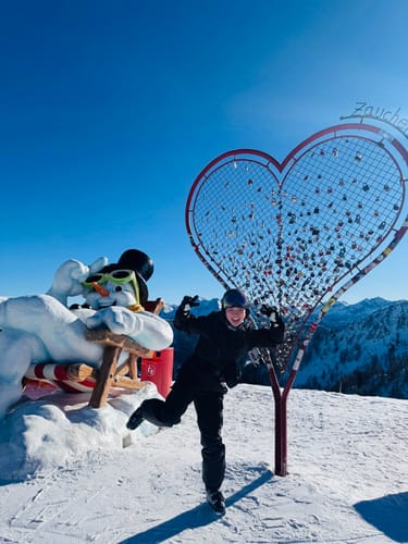 Reviewer posing happily in a dark one-piece ski suit on a sunny, snowy mountain.