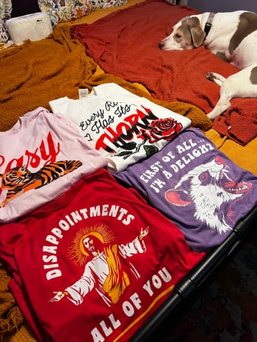 Customer's red Disappointments All Of You Tee shown with a collection of other graphic tees on a bed.