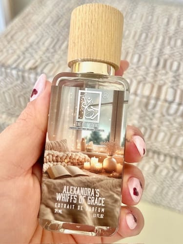 Customer holds the clear glass bottle of Alexandra's Whiffs Of Grace, showing its cozy label.