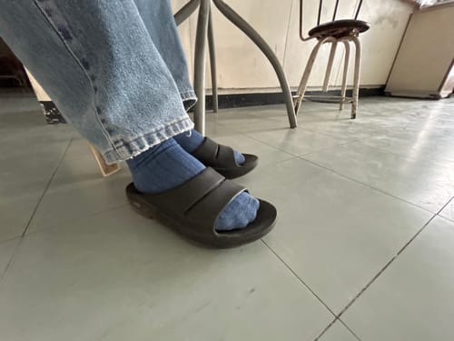 Customer wearing black OOFOS OOAHH Recovery Slides with blue socks and light-wash jeans indoors.