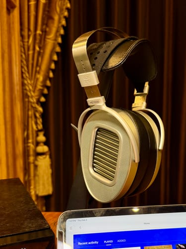 Customer's silver Hifiman HE1000 Unveiled headphones resting on a black stand in a warmly lit room.