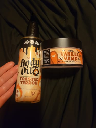 Customer's Toasted Terror Body Oil and another skincare product resting on a dark, rumpled fabric.