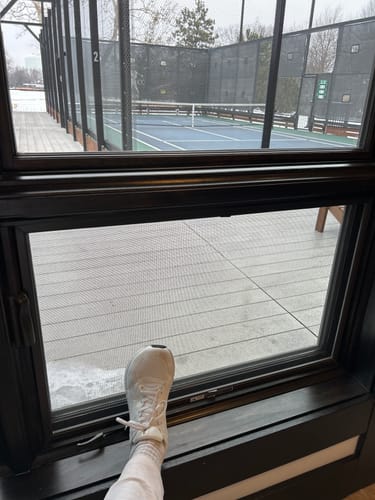 Customer wearing a white Flow Pickleball Shoe, resting their foot on a windowsill overlooking a pickleball court.