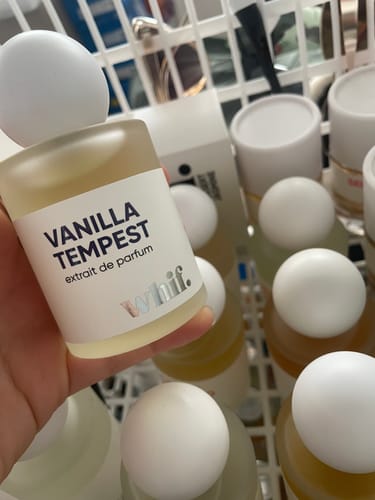 Customer's hand holding a frosted bottle of Vanilla Tempest perfume with a white cap over other similar bottles in a basket.