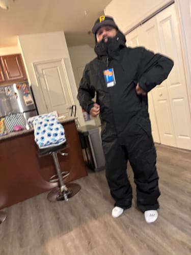 Reviewer standing indoors wearing the new black baggy one-piece ski suit with the hood up.